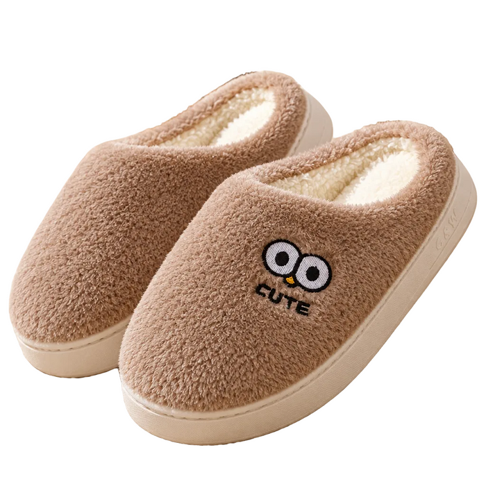 A pair of plush fleece slip-on house slippers in warm taupe brown, shown on a cozy cream rug in a home setting. One slipper features a cute embroidered owl face and the word 'CUTE' on the side.