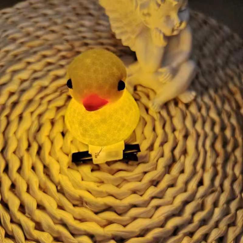 A small yellow duck-shaped night light with a glowing warm yellow body, black eyes, and a red beak, resting on a woven circular mat with a white angel statue in the background.