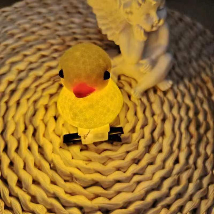 A small yellow duck-shaped night light with a glowing warm yellow body, black eyes, and a red beak, resting on a woven circular mat with a white angel statue in the background.