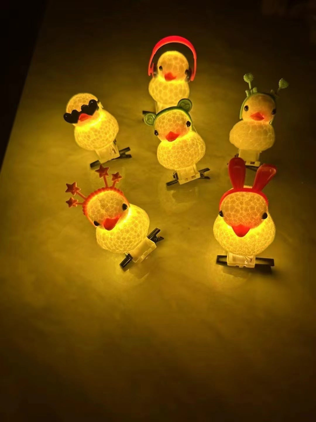 Five small glowing duck-shaped night lights with different cartoon accessories, including a mustache, stars, bunny ears, and headphones, arranged on a dark surface.