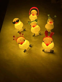 Five small glowing duck-shaped night lights with different cartoon accessories, including a mustache, stars, bunny ears, and headphones, arranged on a dark surface.
