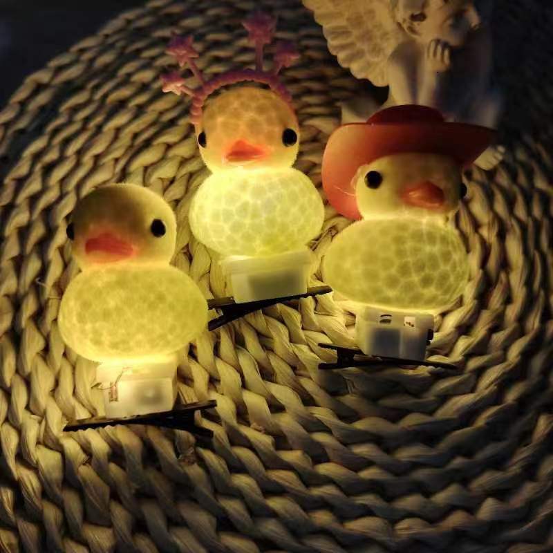 Three small duck-shaped fairy lights glowing on a woven mat, with two wearing hats and one plain, set against a dark background.