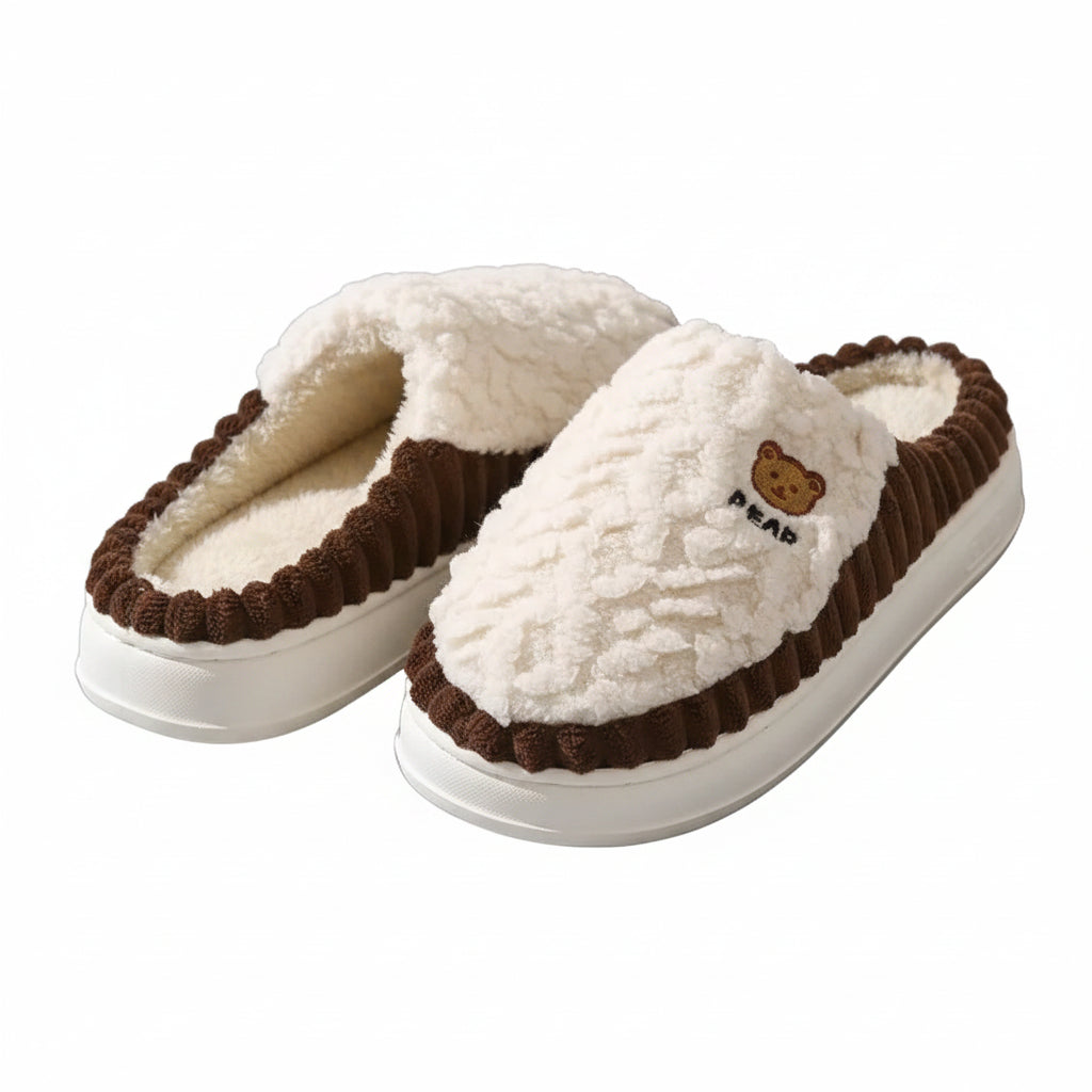 A pair of plush slip-on house slippers with a cream-white textured upper, dark brown ruffled trim, white soles, and a small embroidered bear face with the word 'BEAR' on the side, displayed on shiny silver foil.