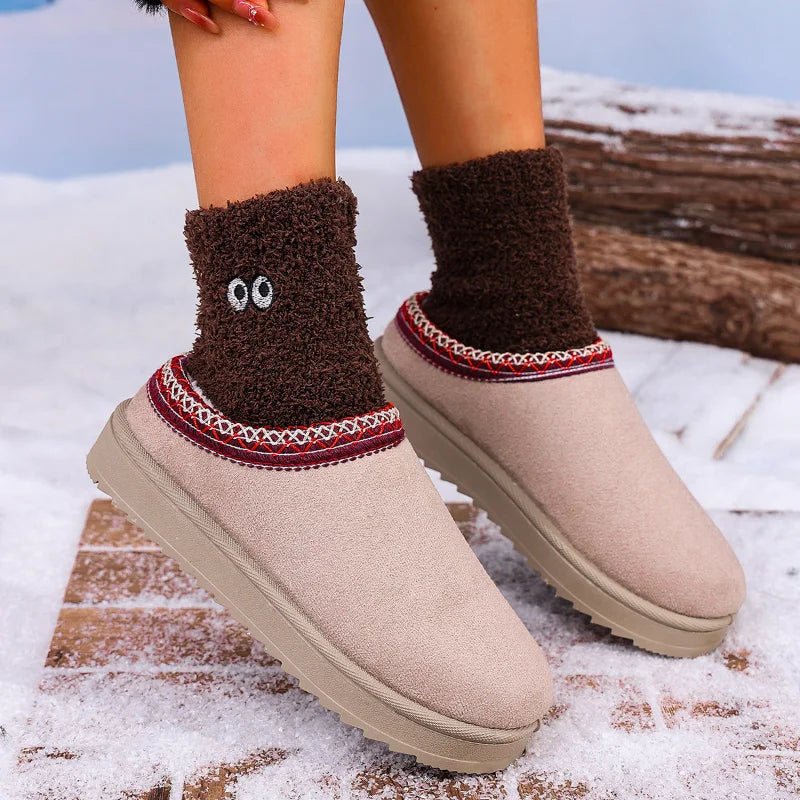 A person wearing beige slip-on winter slippers with brown fuzzy socks featuring embroidered eyes, standing outdoors on snow near a log.