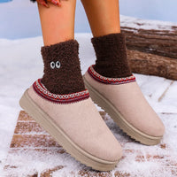 A person wearing beige slip-on winter slippers with brown fuzzy socks featuring embroidered eyes, standing outdoors on snow near a log.