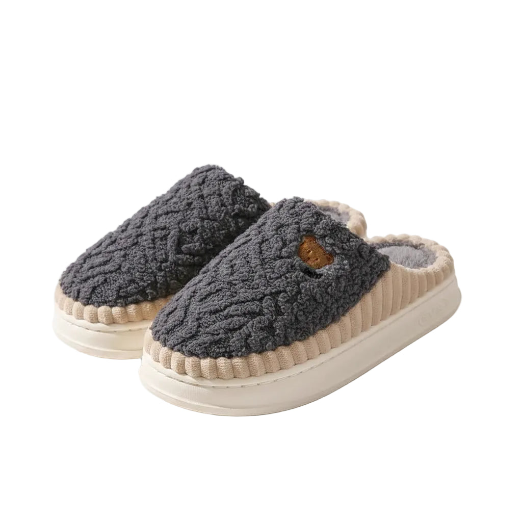 A pair of cozy slip-on slippers with a dark gray cable-knit fleece upper, cream ribbed trim, and a thick cream platform sole, displayed on a wooden floor.