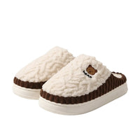 A pair of cozy slip-on house slippers with thick white plush uppers and brown ruffled trim, shown on a wooden floor with warm home decor in the background.