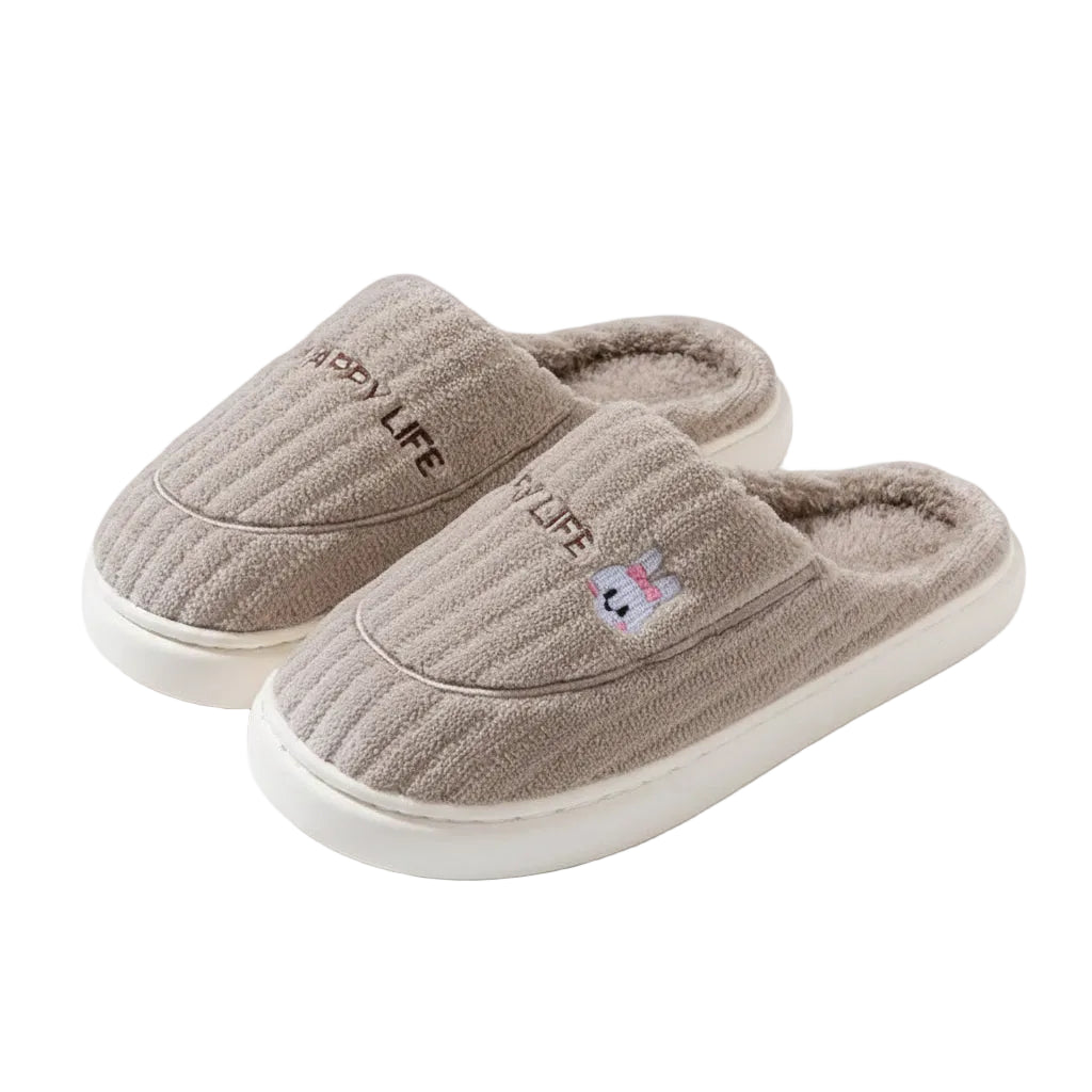 A pair of plush indoor slip-on slippers in a warm taupe-beige color, featuring a ribbed knit exterior, soft fleece lining, embroidered 'HAPPY LIFE' text, and a small cartoon bunny detail. The image includes decorative text overlays and illustrations on a solid taupe background.