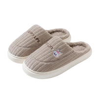 A pair of plush indoor slip-on slippers in a warm taupe-beige color, featuring a ribbed knit exterior, soft fleece lining, embroidered 'HAPPY LIFE' text, and a small cartoon bunny detail. The image includes decorative text overlays and illustrations on a solid taupe background.