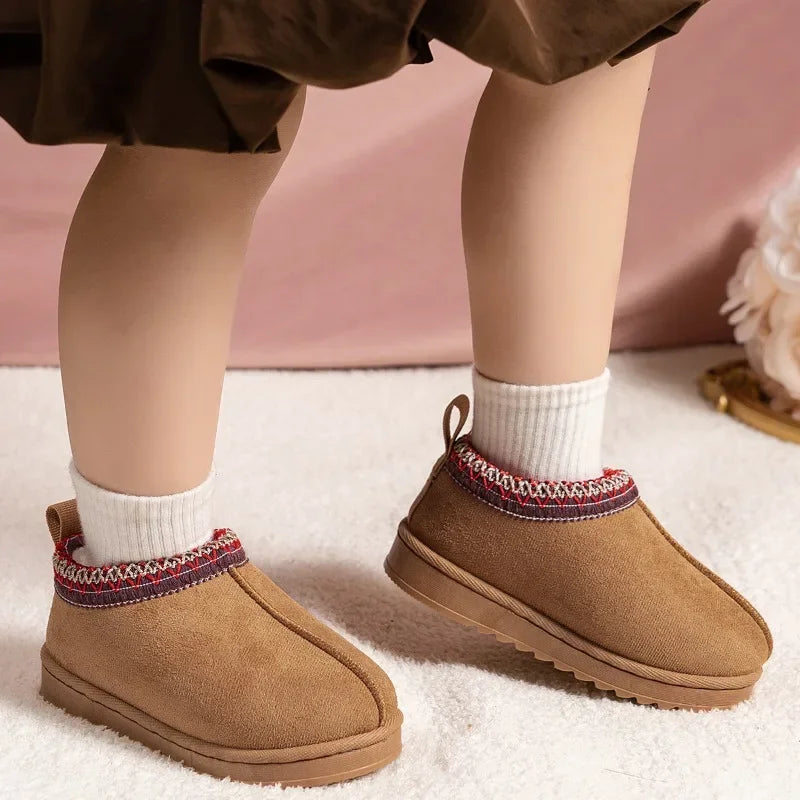 A close-up of a child's legs wearing tan suede-like slipper boots with a colorful knit trim around the collar, paired with white socks, standing on a white textured rug.