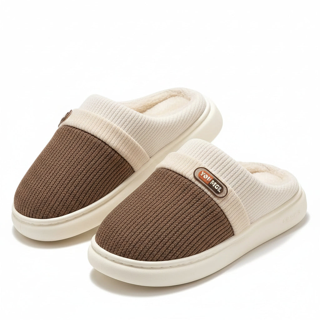 A pair of slip-on indoor slippers with a brown ribbed knit upper, cream ribbed trim, and a cream plush lining, displayed on a wooden stand over a white rug.