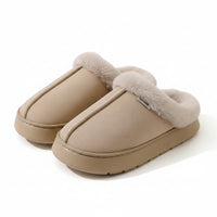 A pair of beige slip-on slippers with fluffy faux-fur trim, displayed on a light beige surface with a white ribbed knit blanket in the background.