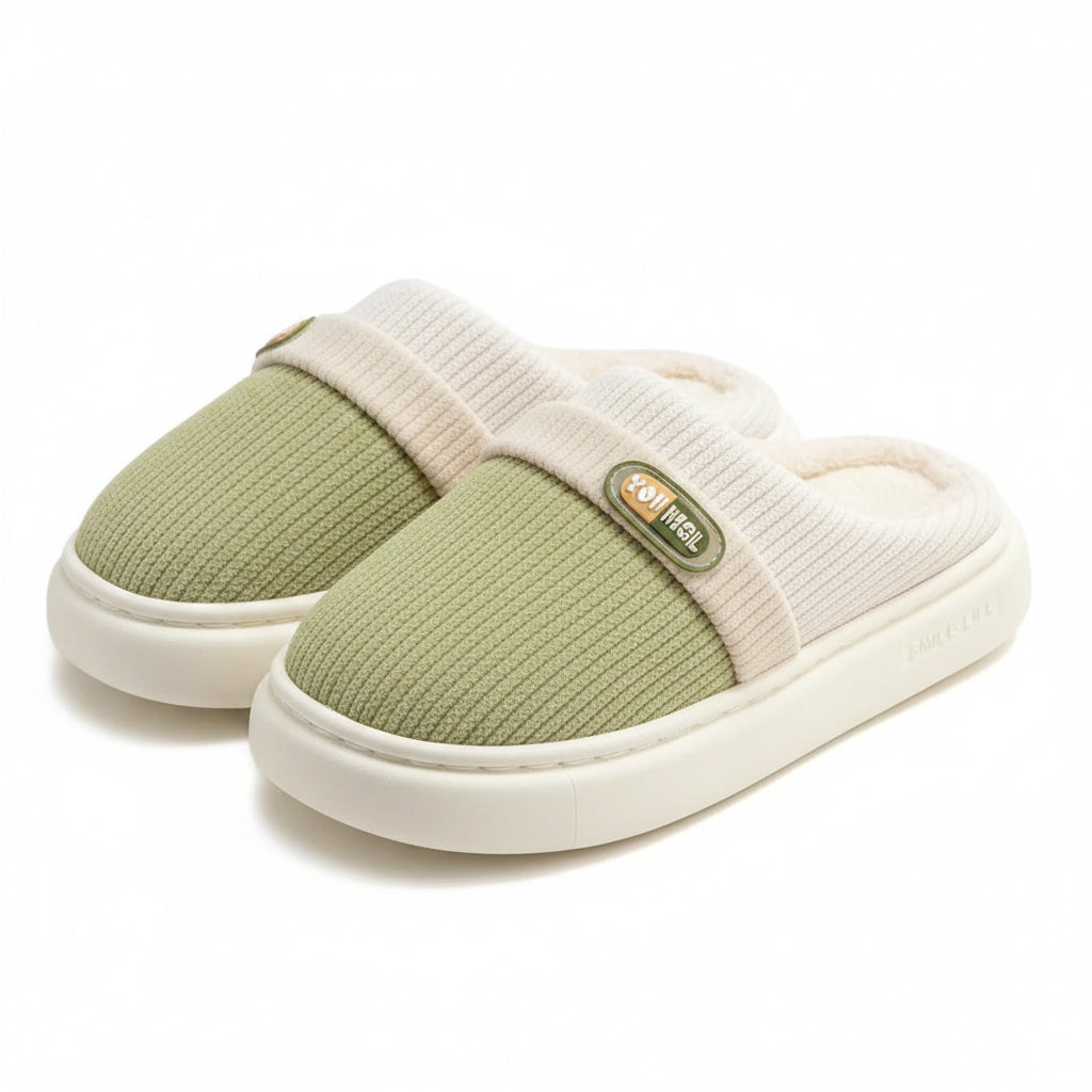 A pair of slip-on indoor slippers with a light green knit upper, cream ribbed band, and cream sherpa lining, shown on a soft rug with wooden furniture in the background.