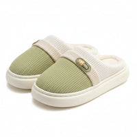 A pair of slip-on indoor slippers with a light green knit upper, cream ribbed band, and cream sherpa lining, shown on a soft rug with wooden furniture in the background.
