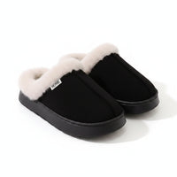 A pair of black slip-on slippers with thick, fluffy cream faux-fur trim around the openings, photographed on a light beige surface with a white ribbed blanket in the background.