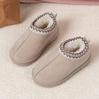 A pair of beige slip-on slippers with a soft white faux-fur lining and a decorative zigzag trim around the collar, displayed on a textured cream carpet.