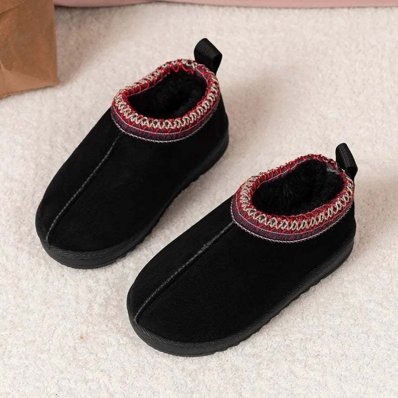 A pair of black slip-on winter boots with a soft fuzzy lining and a red, white, and black patterned trim around the collar, resting on a light beige textured surface.