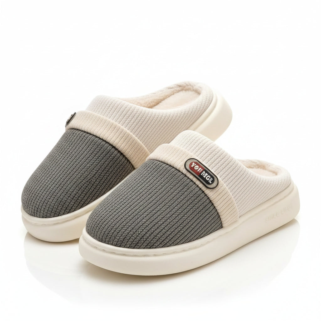 A pair of slip-on indoor slippers with a gray ribbed knit upper, cream ribbed knit collar, cream plush lining, and thick cream soles, displayed on a light rug beside a wooden stand.