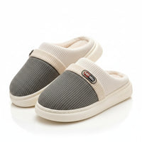 A pair of slip-on indoor slippers with a gray ribbed knit upper, cream ribbed knit collar, cream plush lining, and thick cream soles, displayed on a light rug beside a wooden stand.