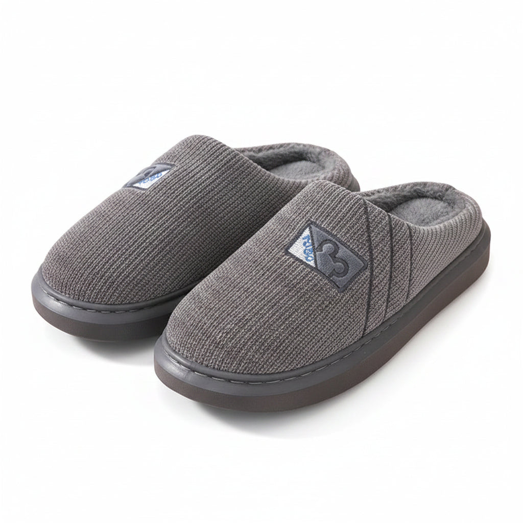 A pair of gray ribbed knit slip-on house slippers with plush gray lining and a small triangular logo patch, displayed on a light wood shelf.