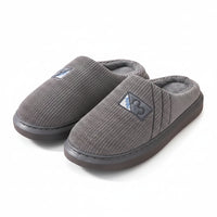 A pair of gray ribbed knit slip-on house slippers with plush gray lining and a small triangular logo patch, displayed on a light wood shelf.