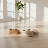 A pair of plush slip-on house slippers in warm beige and tan, shown with one pair on a person's feet and another pair placed on the floor. The slippers have a soft sherpa-like texture, cream lining, and small tan brand tabs.