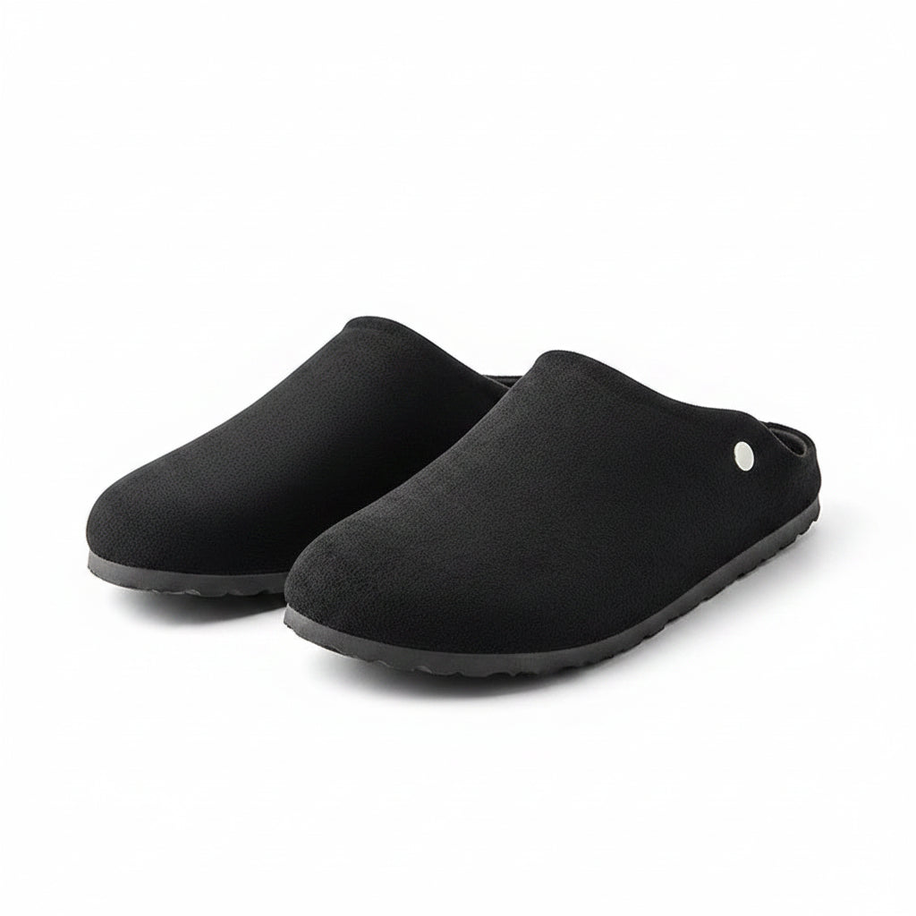 A pair of black slip-on clog-style slippers with a suede-like finish, tan interior lining, and a small silver button detail, displayed on a chrome stand.