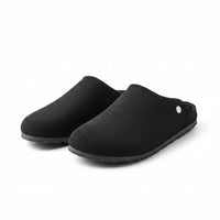 A pair of black slip-on clog-style slippers with a suede-like finish, tan interior lining, and a small silver button detail, displayed on a chrome stand.
