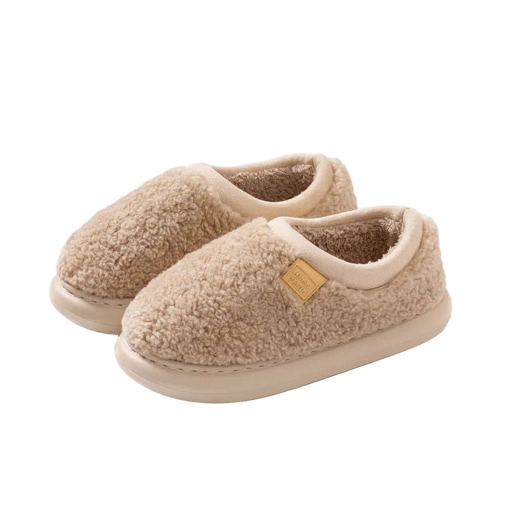 A pair of beige sherpa fleece slip-on slippers with cream trim and a small tan brand patch, shown on a solid light beige background.