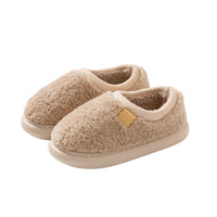 A pair of beige sherpa fleece slip-on slippers with cream trim and a small tan brand patch, shown on a solid light beige background.