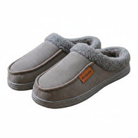 A pair of gray fleece-lined slip-on house slippers with a small orange 'Pimoisie' brand tag, displayed on a wooden surface next to a blue storage box.