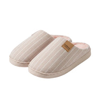 A person holds a pair of plush indoor slippers with a pale pink and white vertical stripe pattern. The slippers have a soft pink lining and a tan leather-like patch on the side with the word 'HOME' printed on it.