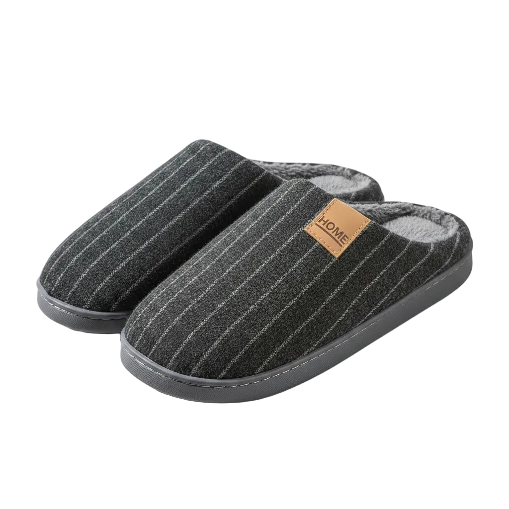 A person holds a pair of dark gray pinstriped indoor slippers with a soft fleece lining. The slippers have a small tan leather patch on the side with the word 'HOME' printed on it.