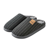 A person holds a pair of dark gray pinstriped indoor slippers with a soft fleece lining. The slippers have a small tan leather patch on the side with the word 'HOME' printed on it.