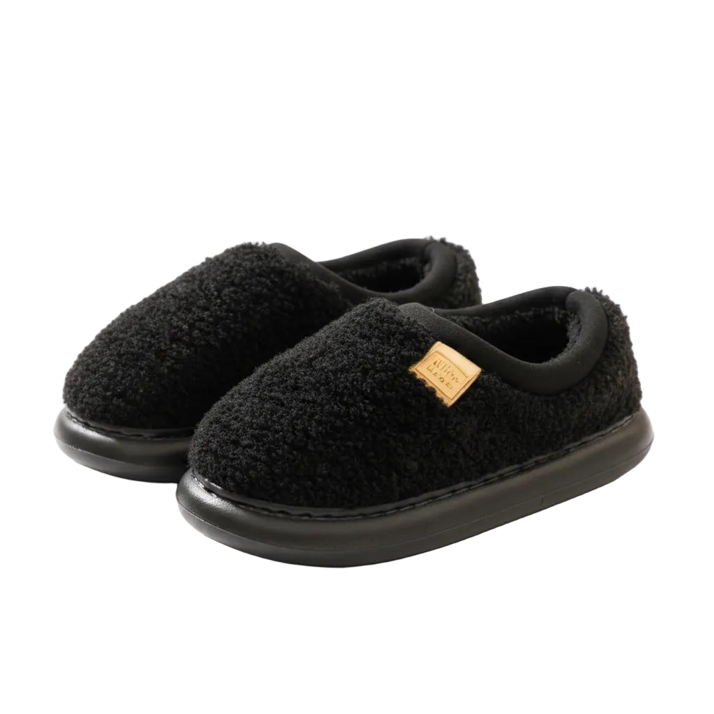 A pair of black fuzzy slip-on house slippers with a small gold brand tag, resting on a cream-colored rug in a warm indoor setting.