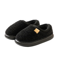 A pair of black fuzzy slip-on house slippers with a small gold brand tag, resting on a cream-colored rug in a warm indoor setting.