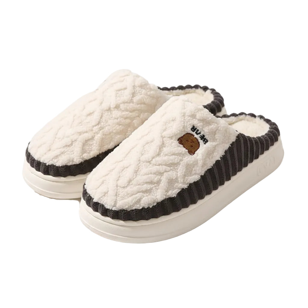 A pair of cozy slip-on slippers with a white textured faux-fur upper, cream sherpa lining, and dark charcoal gray ruffled trim, displayed on a wooden floor.