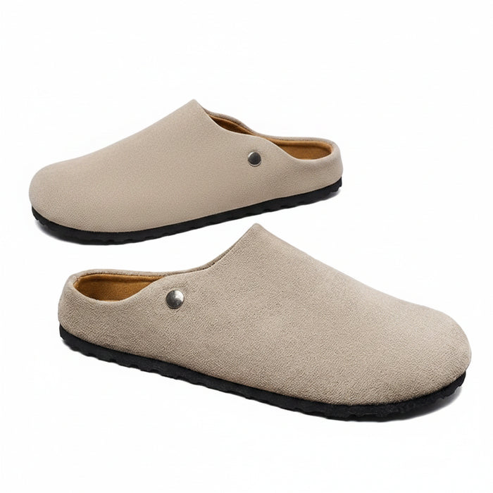A pair of beige slip-on clog-style shoes with black soles, displayed on and around a white shoebox. One shoe is held in a hand wearing a black and gold watch.