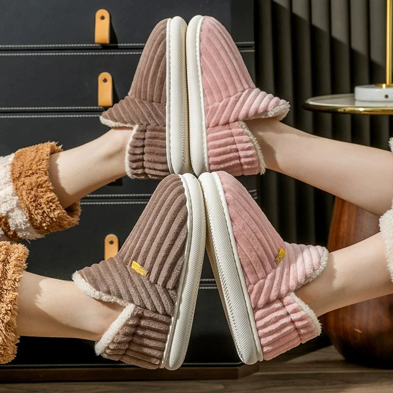 A pair of plush ribbed slipper shoes shown on feet, with one in dusty pink and one in taupe-brown, both with white sherpa lining and white soles. The feet are posed in front of dark drawers in an indoor setting.