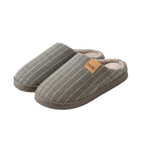A person holds a pair of gray pinstriped fleece slip-on house slippers with a soft brown lining and a tan leather patch that reads 'HOME'.