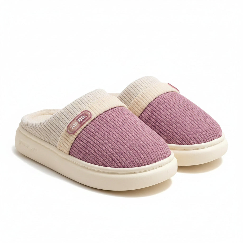 A pair of cozy slip-on indoor slippers with a ribbed mauve-pink upper, cream trim, and plush cream lining, displayed on a wooden stand over a soft rug.