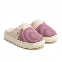 A pair of cozy slip-on indoor slippers with a ribbed mauve-pink upper, cream trim, and plush cream lining, displayed on a wooden stand over a soft rug.