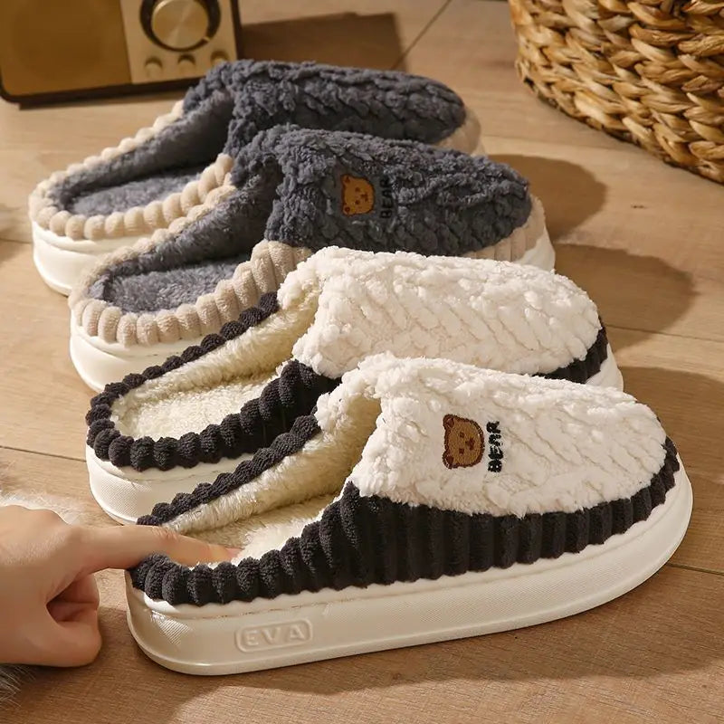 Four plush slip-on slippers arranged on a wooden floor, each with a different color combination: dark gray, cream, black and cream, and white and cream. A small embroidered bear face and the text 'EVA' are visible on the front slippers.