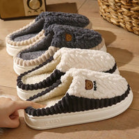 Four plush slip-on slippers arranged on a wooden floor, each with a different color combination: dark gray, cream, black and cream, and white and cream. A small embroidered bear face and the text 'EVA' are visible on the front slippers.