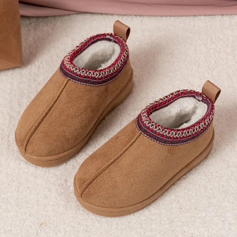 A pair of toddler slip-on winter boots in tan canvas with a white faux-fur lining and a red, white, and navy patterned trim around the collar, shown on a light beige textured surface.