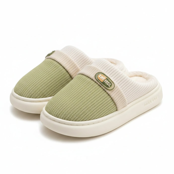 A pair of slip-on indoor slippers with a light sage green knit upper, cream ribbed trim, and a plush cream lining, displayed on a wooden stand over a soft rug. The brand name 'Txlcdn' is visible as a watermark in the top left corner.