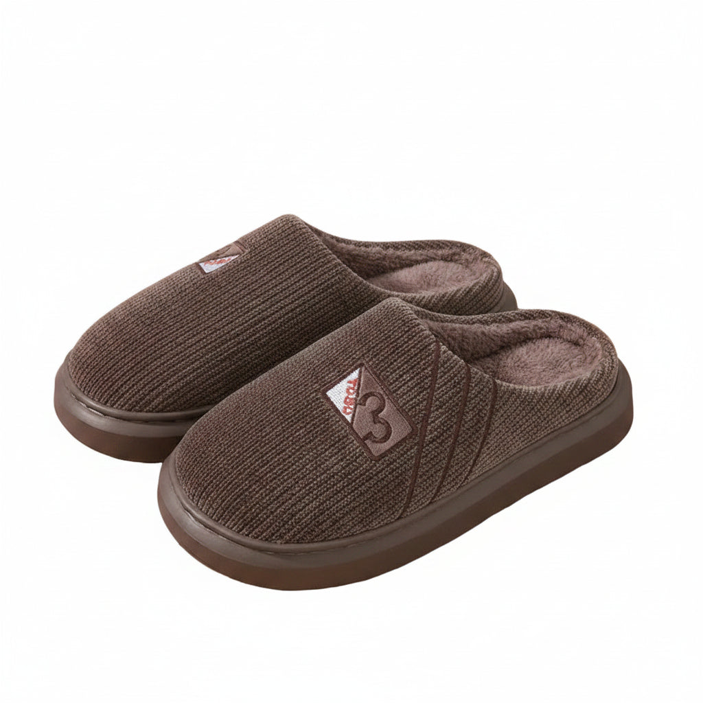 A pair of brown knit slip-on house slippers with plush lining, displayed on a light wood surface next to a white bottle and a patterned bag.
