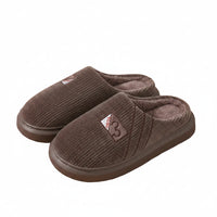 A pair of brown knit slip-on house slippers with plush lining, displayed on a light wood surface next to a white bottle and a patterned bag.