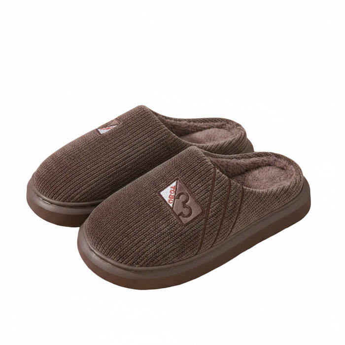 A pair of brown knit slip-on house slippers with plush lining, displayed on a light wood surface next to a white bottle and a patterned bag.