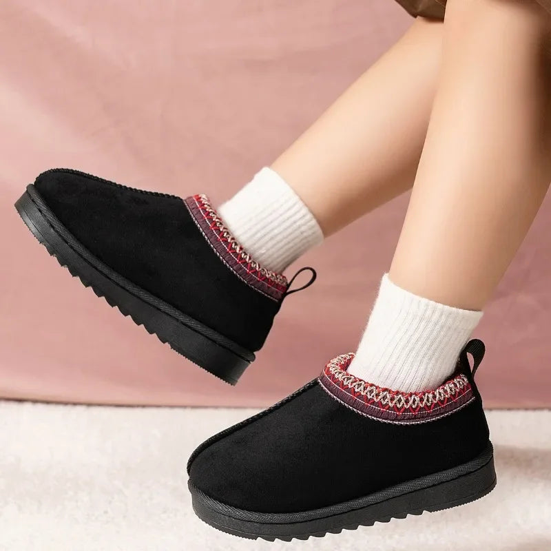 A person wearing black slip-on winter slippers with a red and white patterned trim, paired with white socks, shown against a soft pink backdrop.